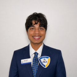 A person with short, curly dark hair smiles at the camera, wearing a blue suit, patterned tie, and a badge and patch labeled FBLA on their jacket, standing against a plain white background.
