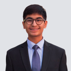 A young person with short dark hair and glasses, wearing a black suit, light blue shirt, and patterned tie, smiles at the camera against a plain light background.