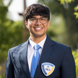 A young man wearing glasses and a blue suit with an FBLA patch on the jacket smiles outdoors; greenery and sunlight are visible in the background.