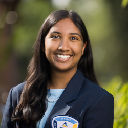 A young woman with long dark hair, wearing a navy blazer with a school emblem, a blue top, and a pearl necklace, smiles outdoors with greenery blurred in the background.