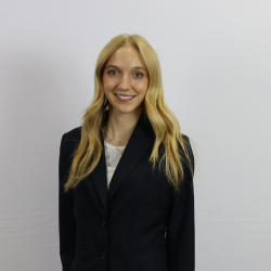 A woman with long blonde hair, wearing a black blazer over a white top, stands in front of a plain white background, smiling at the camera.