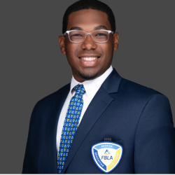A young man in a navy suit, white shirt, and patterned blue tie smiles at the camera. He wears glasses and an FBLA badge on his blazer against a plain gray background.