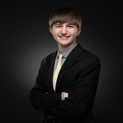 A young person with straight brown hair, wearing a black suit, gray shirt, and yellow tie, stands smiling with arms crossed against a dark, gradient background.