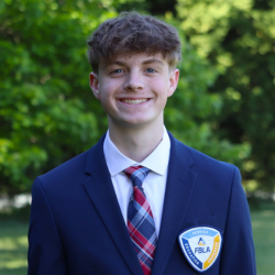 A young person with light brown hair, wearing a navy suit, white shirt, and plaid tie, smiles outdoors. They have an FBLA insignia on their jacket. Green trees and grass are visible in the background.