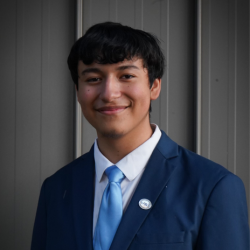 A young man with short dark hair wearing a blue suit, white shirt, and light blue tie stands in front of a gray paneled wall, smiling at the camera.