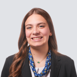 A young woman with long brown hair smiles at the camera. She is wearing a black blazer, a white top, and a blue patterned scarf. The background is light gray.