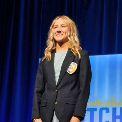A woman with wavy blonde hair, wearing a light blue dress and a dark blazer with a yellow name badge, stands and smiles on a stage with a dark blue curtain and a blue and yellow sign in the background.