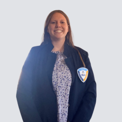 A woman with long hair smiles at the camera, wearing a navy blazer with a blue and orange badge and a blue patterned blouse, standing against a plain light gray background.
