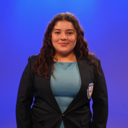 A young woman with long curly hair, wearing a blue dress and a dark blazer with a badge, stands smiling in front of a blue gradient background.