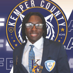 A young man in a suit and tie stands smiling in front of a large Kemper County school logo. He wears glasses and a badge with the FBLA emblem on his jacket.