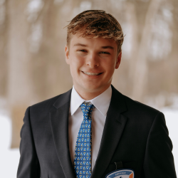 A young man wearing a dark suit, white shirt, and patterned blue tie smiles at the camera outdoors on a snowy day. He has short, light brown hair and is wearing a name badge.