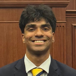 A young man with dark hair, wearing a dark suit, white shirt, and yellow striped tie, smiles at the camera while standing in front of a wooden panel background.