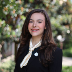 A young woman with long brown hair, wearing a black blazer, a white scarf, and a pin, smiles while standing outdoors in a garden setting with greenery and blurred trees in the background.