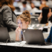 Two young people sit at a table, focused on their laptops in a busy conference or classroom setting, surrounded by others working on computers.